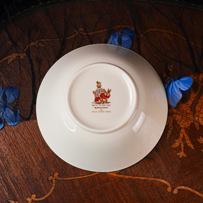 May include: A white ceramic saucer featuring a central stamp with a crown, a group of rabbits, and the text English Fine Bone China Bunnykins Regd. Trade Mark. The underside of the saucer is smooth and circular with a raised foot rim.