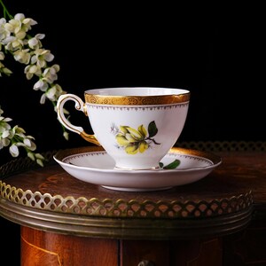 Peut inclure: Une tasse et une soucoupe blanches avec des bordures dor&eacute;es et un motif floral jaune. La tasse a une anse incurv&eacute;e et repose sur une soucoupe assortie. L'ensemble est pos&eacute; sur une surface en bois.