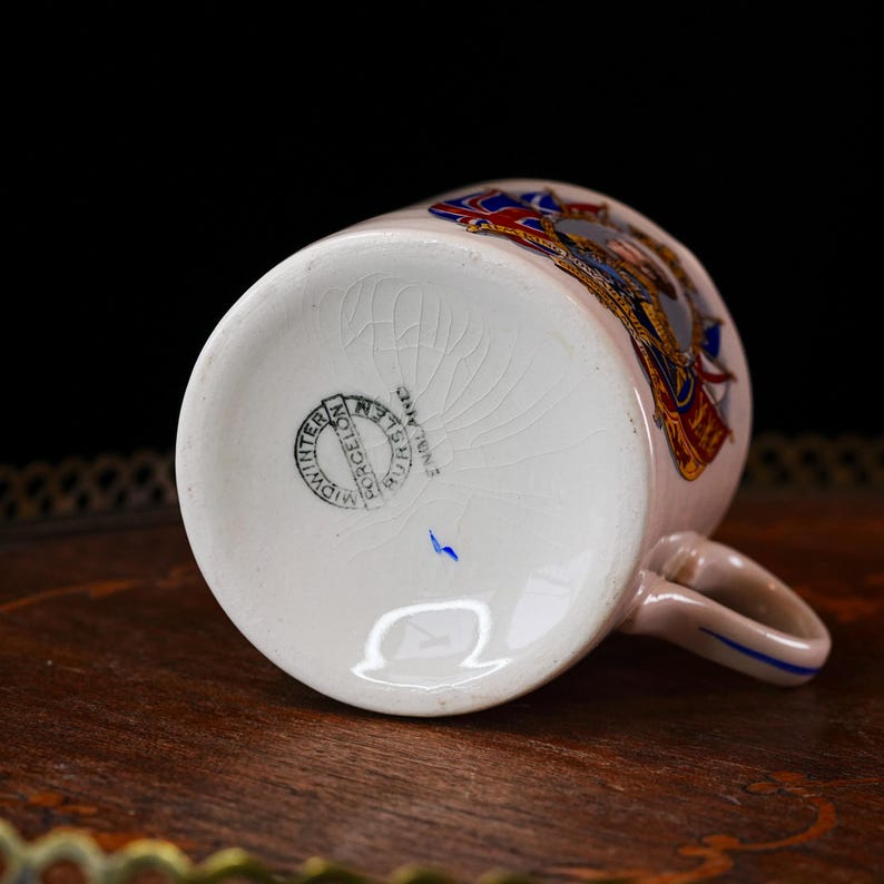 Op de afbeelding: Cr&egrave;mekleurige porseleinen mok met een handvat, met een kleurrijk wapenontwerp. De mok heeft een blauw accent op het handvat en een stempel met de tekst "Midwinter, Burslem, England". De mok staat op een houten oppervlak.