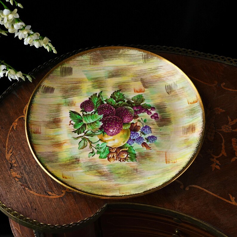 May include: A round decorative plate with a gold rim and a painted fruit design. The plate features a central image of purple berries, green leaves, and a yellow fruit. The background has a textured, light green and gold pattern.