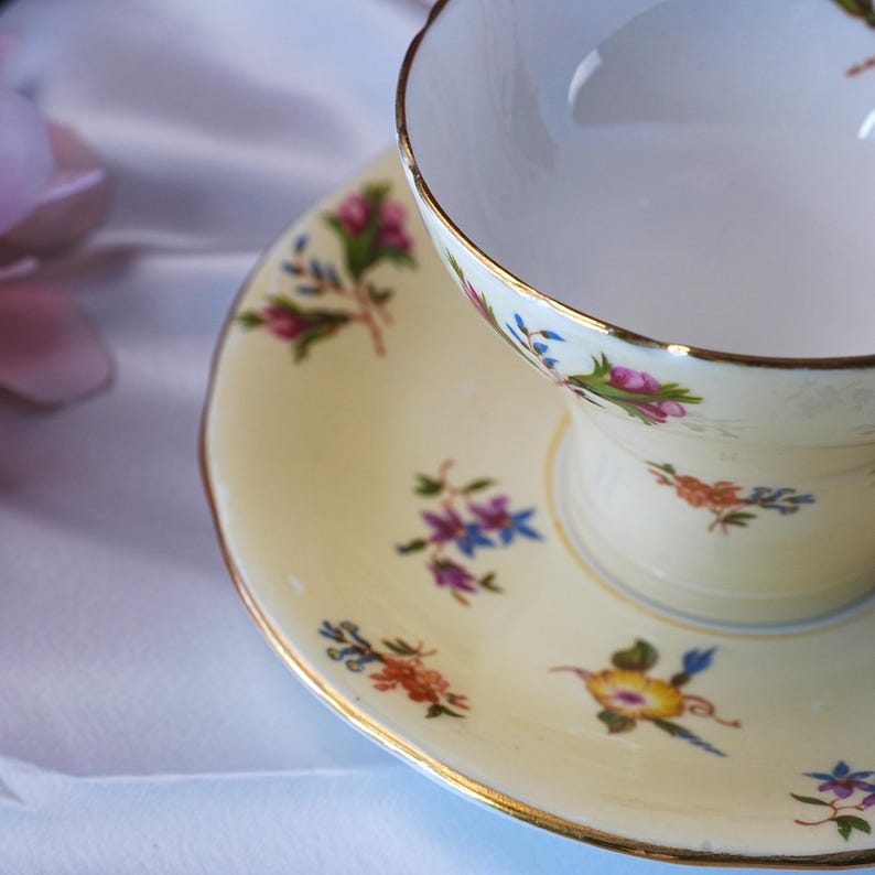 Op de afbeelding: Een vintage theekop en schotel set. De kop en schotel zijn cr&egrave;mekleurig met gouden randen en bloemmotieven in roze, blauw en geel. De theekop is leeg en staat op de bijpassende schotel.