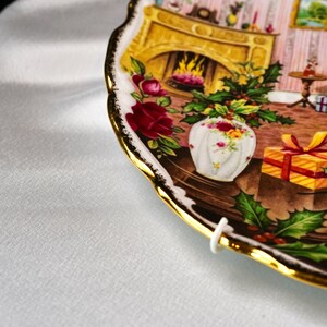Pu&ograve; includere: Un piatto decorativo con bordo dorato e una dettagliata scena natalizia. Il piatto presenta un camino, un vaso di fiori e un regalo avvolto. Il bordo del piatto ha un design smerlato.