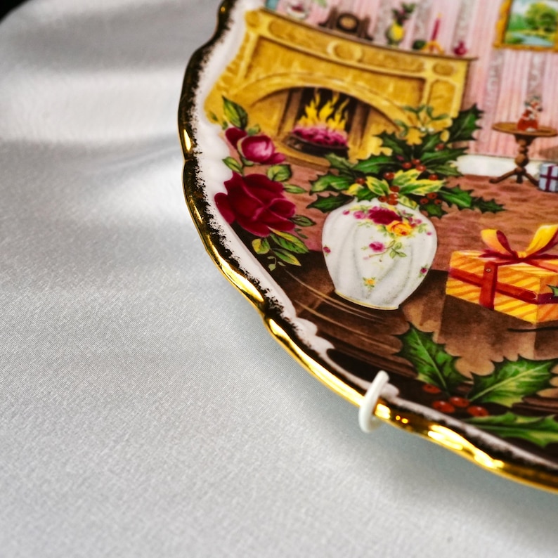 Pu&ograve; includere: Un piatto decorativo con bordo dorato e una dettagliata scena natalizia. Il piatto presenta un camino, un vaso di fiori e un regalo avvolto. Il bordo del piatto ha un design smerlato.