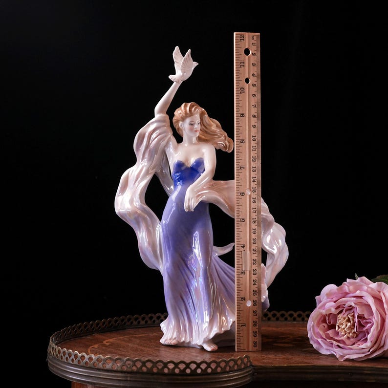 May include: A porcelain figurine of a woman in a flowing purple gown, holding a white dove. The figurine is approximately 25 cm tall. The woman has long, flowing auburn hair and is draped in a light, iridescent fabric.