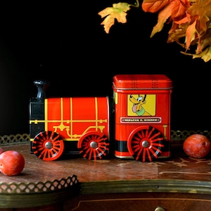 Peut inclure: Un train jouet rouge et jaune avec des roues noires. Le train présente un wagon rectangulaire avec un dessin de Pluto et le texte "WALTER E. DISNEY". Le train est en métal et repose sur une surface en bois.