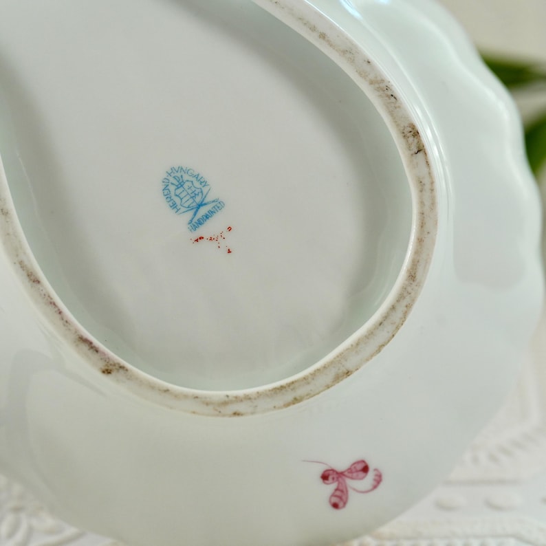 May include: A white porcelain serving dish with a scalloped edge. The dish has a blue and red hand-painted emblem and a small pink floral design. The rim has a brown, aged appearance.