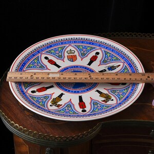 Puede incluir: Un plato conmemorativo de cer&aacute;mica blanca con un borde azul y el texto SILVER JUBILEE 1952-1977 GOD SAVE OUR GRACIOUS QUEEN. El centro muestra un escudo real, guardias y motivos florales. Una regla muestra un di&aacute;metro de 26,7 cent&iacute;metros.