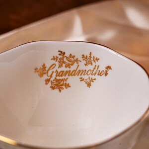 K&ouml;nnte beinhalten: Eine wei&szlig;e Keramik-Sauciere mit goldenem Rand und dem Wort "Grandmother" in goldener Schrift, umgeben von floralen Designs. Das Boot hat eine klassische, elegante Form.
