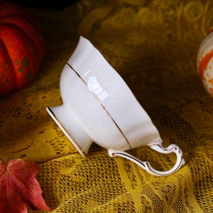 May include: A white porcelain teacup with a gold trim and handle. The teacup has a unique, petal-like shape and sits on a small pedestal base. The handle is also detailed with gold accents. The teacup is set against a yellow lace tablecloth.