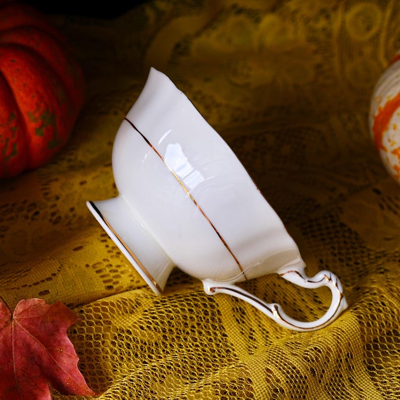 May include: A white porcelain teacup with a gold trim and handle. The teacup has a unique, petal-like shape and sits on a small pedestal base. The handle is also detailed with gold accents. The teacup is set against a yellow lace tablecloth.