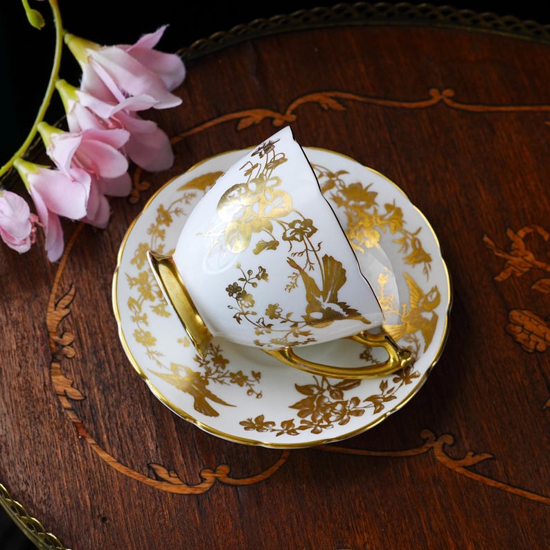 Op de afbeelding: Een witte kop en schotel met gouden bloemen- en vogelontwerpen. De kop heeft een gouden oor en rand, passend bij het ontwerp van de schotel. De set staat op een donker houten oppervlak, met roze bloemen op de achtergrond.