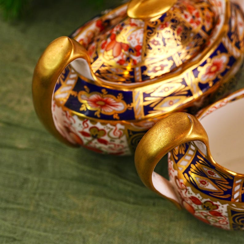 May include: Close-up of a decorative porcelain tea set. The set features a gold handle and rim, with a detailed pattern of red, blue, and gold floral designs. The background is a muted green fabric.