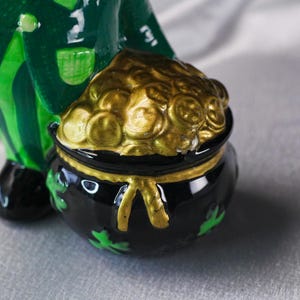 May include: A ceramic leprechaun figurine holding a black pot filled with gold coins. The pot has a gold rim and shamrock details. The leprechaun wears a green coat and striped pants.