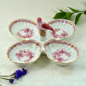 May include: A white porcelain serving dish with four scalloped sections, each adorned with pink floral patterns and gold trim. A pink and red bird figurine perches atop the center, adding a decorative touch to this elegant piece.