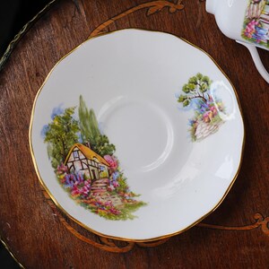 May include: White porcelain saucer with a gold rim, featuring a colorful cottage scene with a yellow roof, surrounded by flowers and trees. The saucer has a scalloped edge, and is part of a tea set.