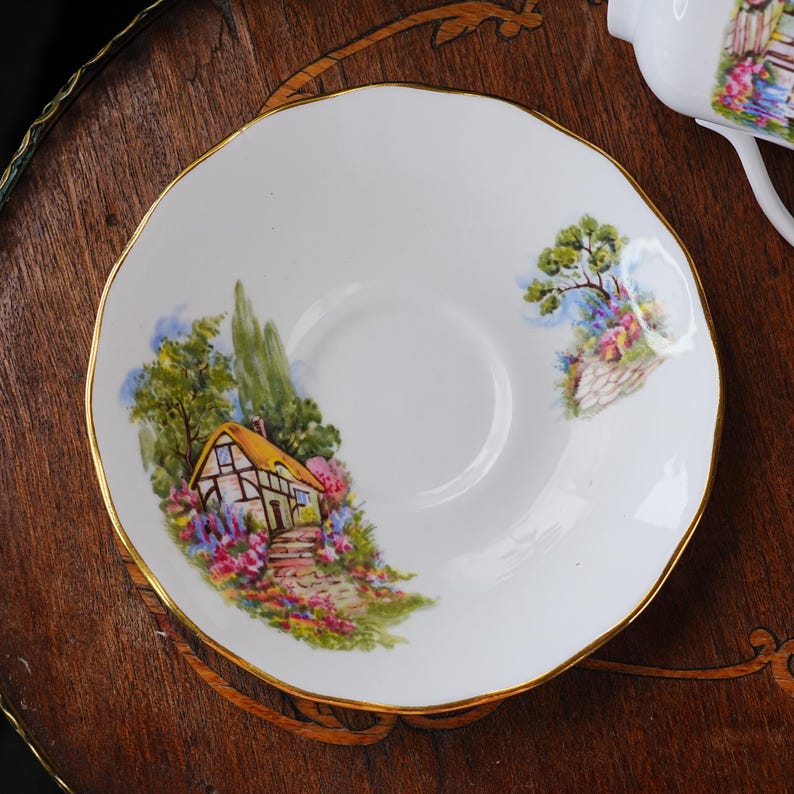 May include: White porcelain saucer with a gold rim, featuring a colorful cottage scene with a yellow roof, surrounded by flowers and trees. The saucer has a scalloped edge, and is part of a tea set.