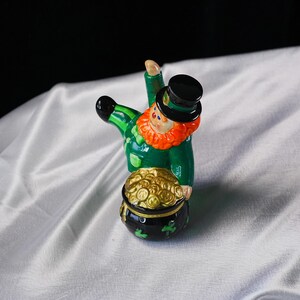May include: A ceramic leprechaun figurine with a green coat, black top hat, and orange beard. The leprechaun is holding a pot of gold coins. The pot is black with green shamrocks. A festive St. Patrick's Day decoration.
