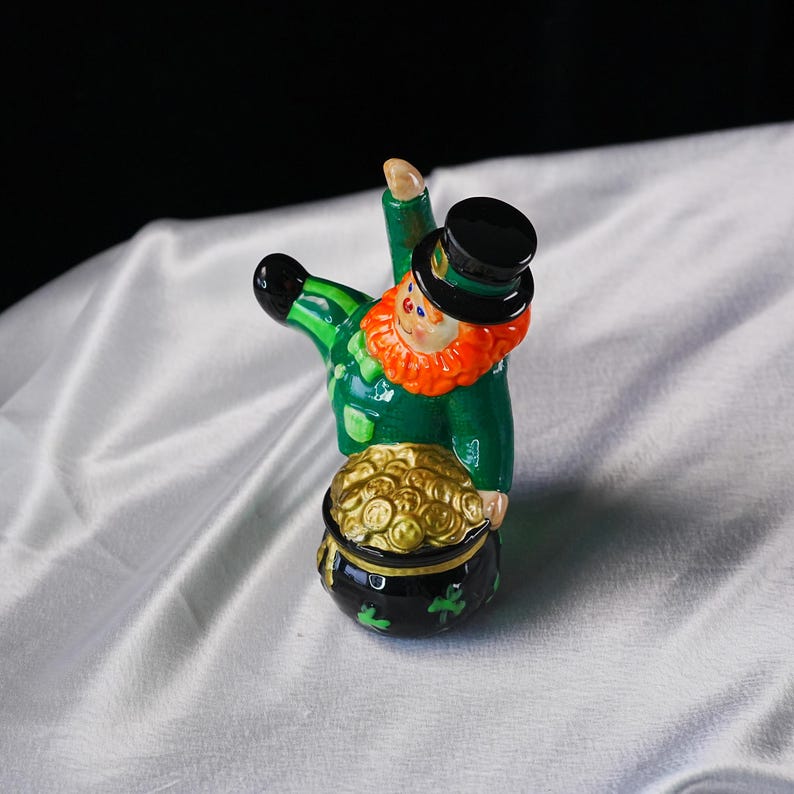 May include: A ceramic leprechaun figurine with a green coat, black top hat, and orange beard. The leprechaun is holding a pot of gold coins. The pot is black with green shamrocks. A festive St. Patrick's Day decoration.