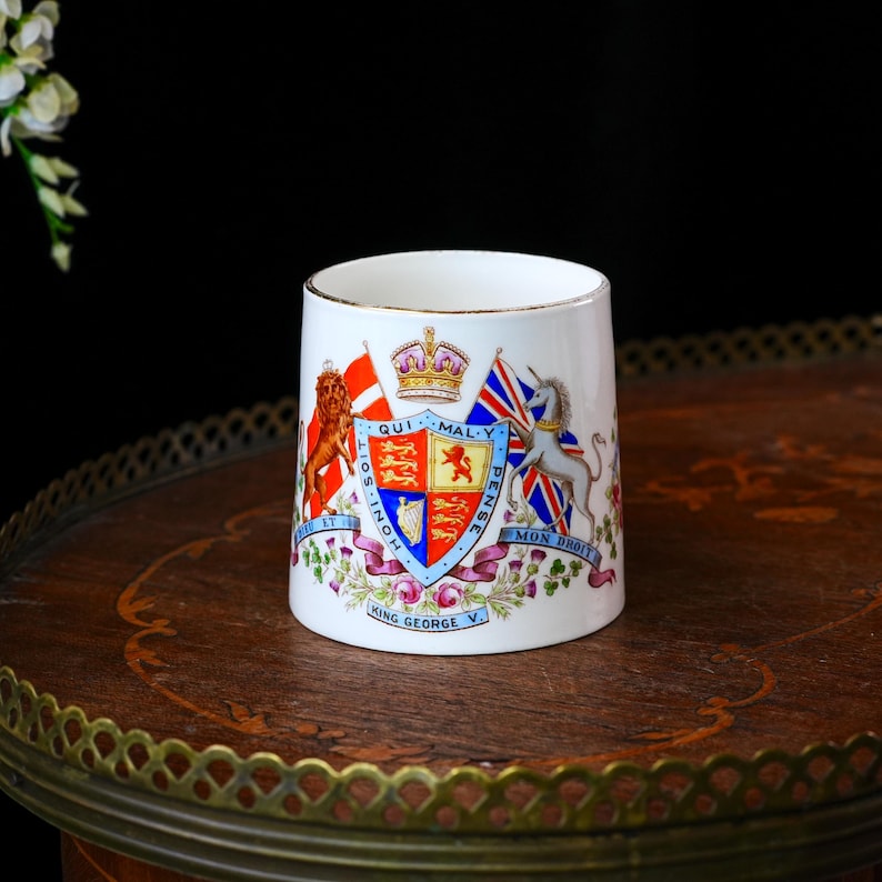 Op de afbeelding: Een witte keramische mok met een gouden rand, met een gedetailleerd wapenschild. Het ontwerp omvat vlaggen, een kroon en de tekst "King George V". De mok staat op een donker houten oppervlak.