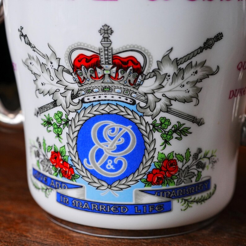 May include: White ceramic mug with a royal crest design. The crest features a red crown, blue emblem, and the words "IN MARRIED LIFE". The mug has a silver handle and is decorated with red roses and silver accents.