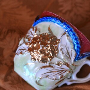 May include: A close-up of a vintage teacup with a floral design. The cup features a gold, white, and light green color scheme with gold accents. The rim is red and blue. The handle is white.