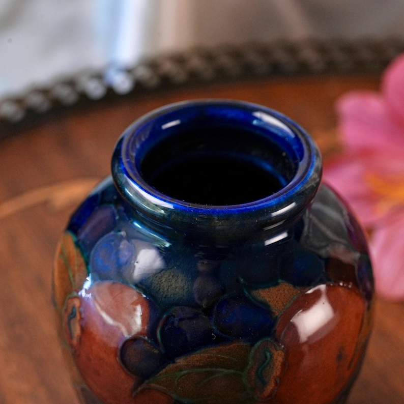 May include: A small ceramic vase with a glossy finish, featuring a deep cobalt blue rim and neck. The body of the vase displays a dark, mottled pattern of rich brown and navy blue tones, with subtle hints of green and orange throughout the surface.