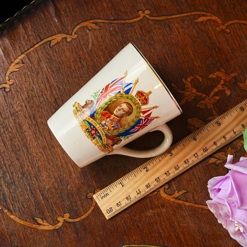 Puede incluir: Taza de cer&aacute;mica color crema con borde dorado, con un retrato del rey Eduardo VII, banderas y texto. La taza mide aproximadamente 10 cm de alto y tiene un asa. Una regla de madera est&aacute; al lado de la taza.