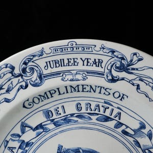 May include: White ceramic plate with blue decorative elements and text. The plate features the words "JUBILEE YEAR" and "COMPLIMENTS OF DEI GRATIA" in a stylized font. The design includes floral and ribbon motifs.