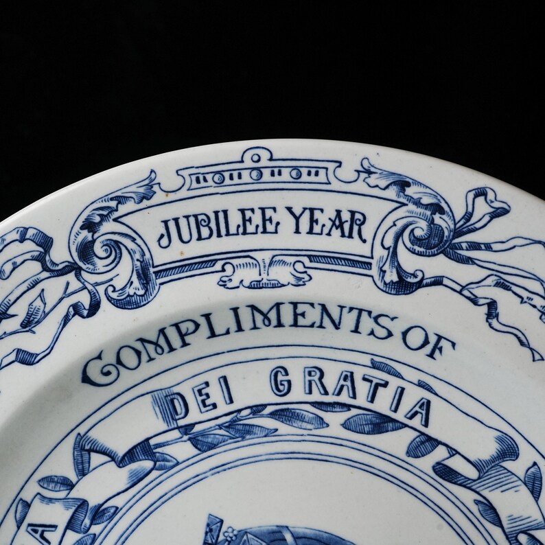 May include: White ceramic plate with blue decorative elements and text. The plate features the words "JUBILEE YEAR" and "COMPLIMENTS OF DEI GRATIA" in a stylized font. The design includes floral and ribbon motifs.