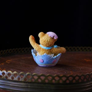May include: A small, tan teddy bear figurine wearing a pink bonnet and blue scarf, seated inside a light blue eggshell-shaped container with pink and purple accents. The bear is waving its right arm.