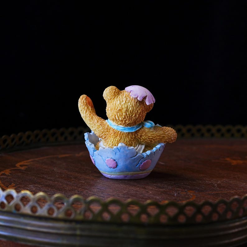 May include: A small, tan teddy bear figurine wearing a pink bonnet and blue scarf, seated inside a light blue eggshell-shaped container with pink and purple accents. The bear is waving its right arm.