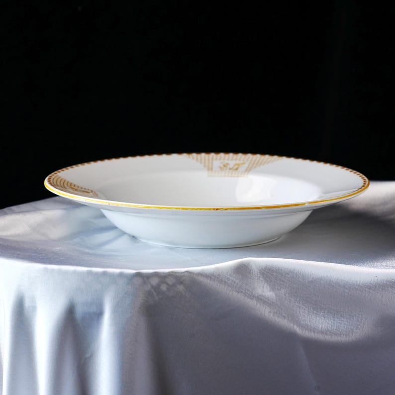 May include: A white ceramic soup bowl with a gold rim and decorative gold accents. The bowl has a wide, shallow design and sits on a white tablecloth. The rim has a scalloped edge.