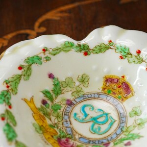 Op de afbeelding: Een klein schaaltje met geschulpte rand en een cr&egrave;mekleurige basis. Het is versierd met een bloemen- en embleemdesign, inclusief een kroon, distels en een monogram. De rand heeft een gouden afwerking.