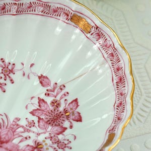 May include: Close-up of a white porcelain dish with a scalloped edge and a gold rim. The dish is decorated with pink floral patterns and accents. The design includes stylized flowers and leaves, creating an elegant aesthetic.