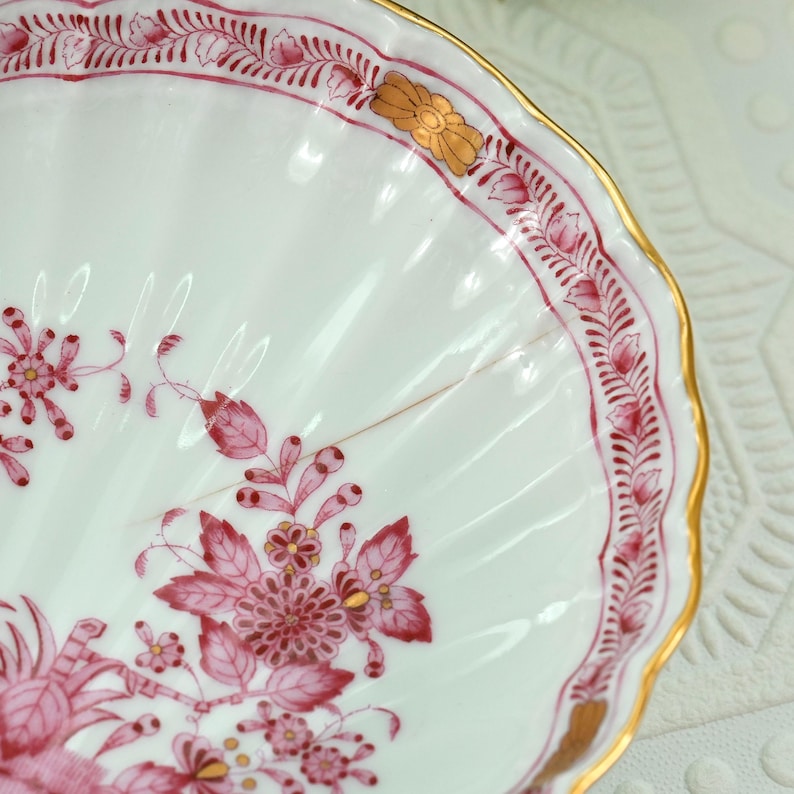 May include: Close-up of a white porcelain dish with a scalloped edge and a gold rim. The dish is decorated with pink floral patterns and accents. The design includes stylized flowers and leaves, creating an elegant aesthetic.
