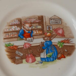 May include: A white ceramic plate featuring a colorful illustration of a pig shopkeeper behind a counter labeled Mr. Piggly's Stores, with rabbits shopping for carrots. The scene includes shelves with jars, a sign, and a sack labeled Piglet Carrots.