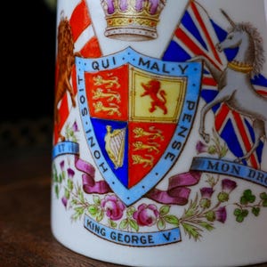 Op de afbeelding: Detailopname van een witte keramische mok met een gedetailleerd koninklijk wapen. Het wapen bevat een schild met verschillende emblemen, vlaggen en de tekst "King George V." Het ontwerp bevat rode, blauwe, gouden en paarse accenten.