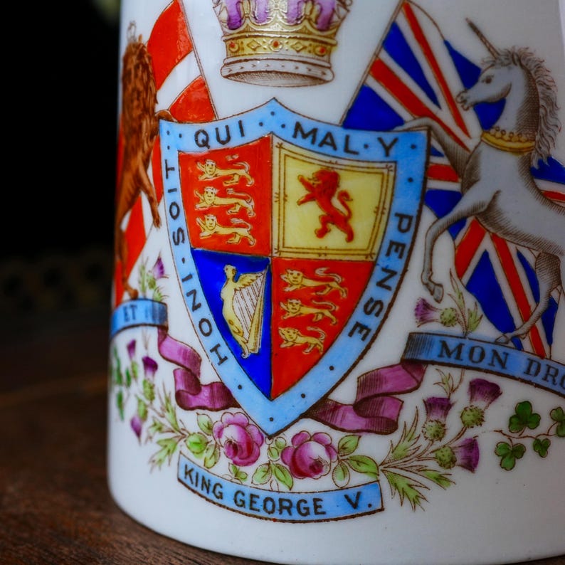 Op de afbeelding: Detailopname van een witte keramische mok met een gedetailleerd koninklijk wapen. Het wapen bevat een schild met verschillende emblemen, vlaggen en de tekst "King George V." Het ontwerp bevat rode, blauwe, gouden en paarse accenten.