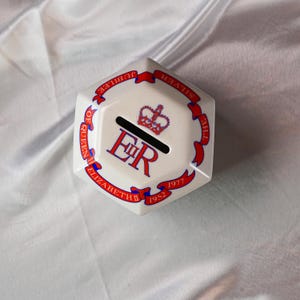 Op de afbeelding: Een witte zeshoekige keramische spaarpot met rode en blauwe accenten. De spaarpot heeft de tekst "SILVER JUBILEE OF QUEEN ELIZABETH 1952-1977" rond de randen, een kroon en de letters "ER".