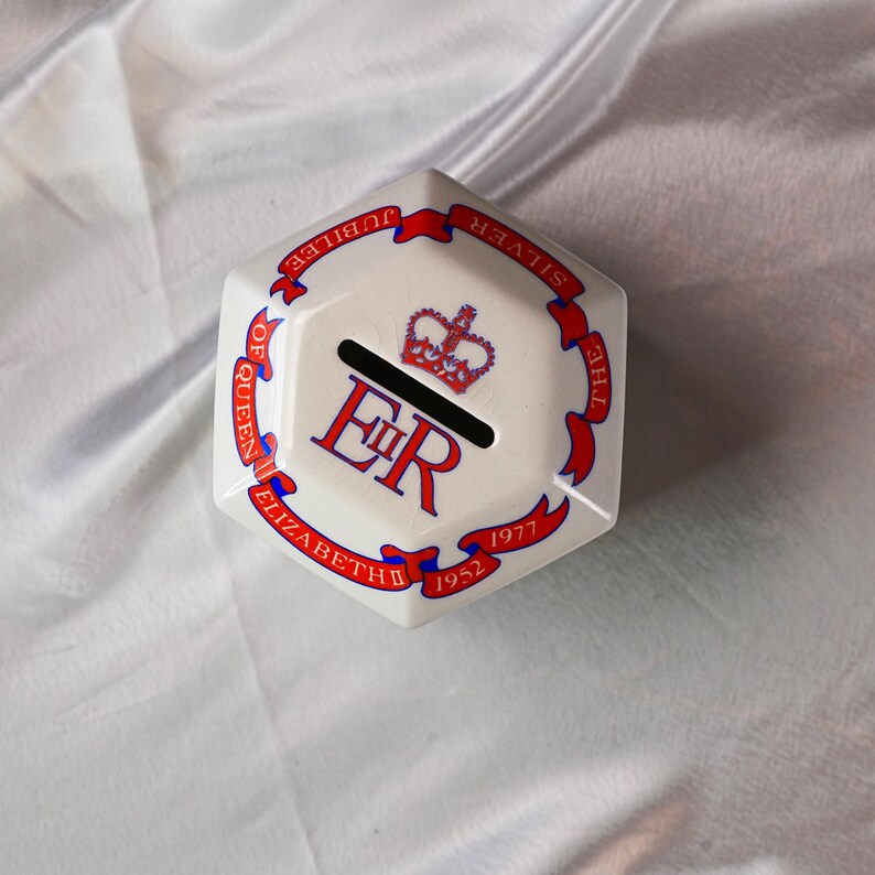 Op de afbeelding: Een witte zeshoekige keramische spaarpot met rode en blauwe accenten. De spaarpot heeft de tekst "SILVER JUBILEE OF QUEEN ELIZABETH 1952-1977" rond de randen, een kroon en de letters "ER".