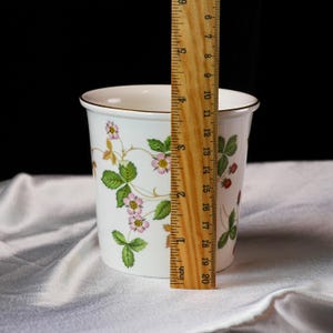 Op de afbeelding: Witte keramische container met een gouden rand, versierd met een aardbeienplantontwerp met groene bladeren, roze bloemen en rode bessen. De container is ongeveer 15 cm hoog.