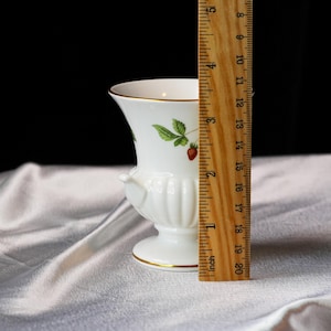 Op de afbeelding: Een witte keramische vaas met een aardbeienontwerp, met groene bladeren en rode bessen. De vaas heeft een gouden rand en voet en is ongeveer 13 cm hoog.