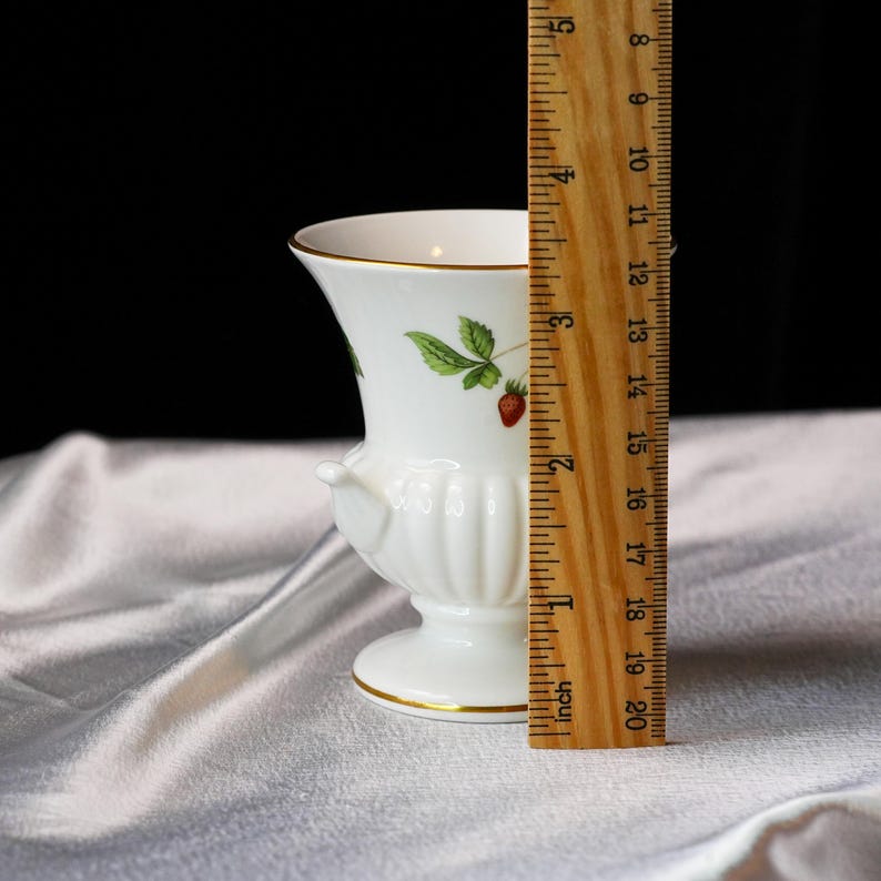 Op de afbeelding: Een witte keramische vaas met een aardbeienontwerp, met groene bladeren en rode bessen. De vaas heeft een gouden rand en voet en is ongeveer 13 cm hoog.