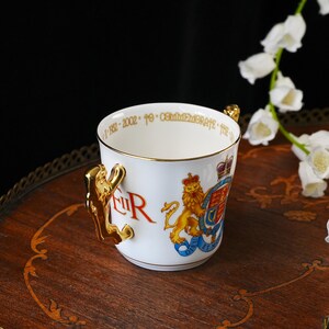 Puede incluir: Una taza conmemorativa de porcelana blanca con un asa dorada en forma de le&oacute;n. El exterior muestra el escudo real, el monograma EIIR en rojo y un texto dorado en el borde interior que dice 1952-2002 To Commemorate the Golden Jubilee.
