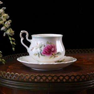 May include: A white teacup and saucer set featuring a pink rose floral pattern. The teacup has a curved handle with gold detailing. The saucer is round with a scalloped edge. The set is displayed on a dark wooden surface.