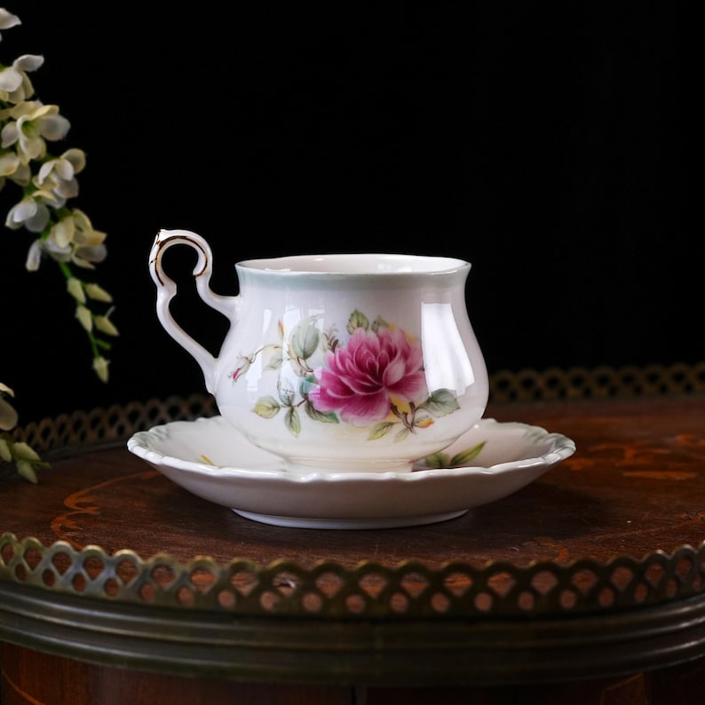 May include: A white teacup and saucer set featuring a pink rose floral pattern. The teacup has a curved handle with gold detailing. The saucer is round with a scalloped edge. The set is displayed on a dark wooden surface.