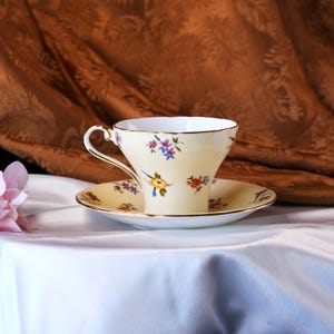 Op de afbeelding: Een vintage theekop en schotel set. De kop en schotel zijn cr&egrave;mekleurig met een gouden rand en delicate bloempatronen in blauw, geel en rood. De set staat op een wit oppervlak met een bruine achtergrond.