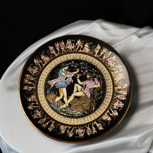 May include: A decorative black plate with a gold rim and a detailed illustration of figures in classical attire. The central image depicts two figures, one with a bow and arrow, and the other reclining. The plate's border features additional figures.