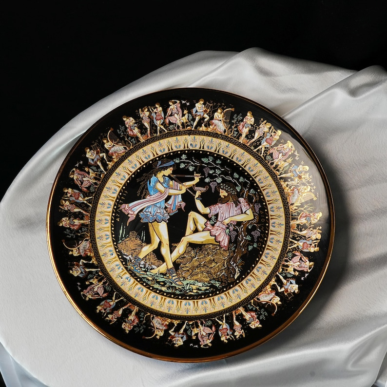 May include: A decorative black plate with a gold rim and a detailed illustration of figures in classical attire. The central image depicts two figures, one with a bow and arrow, and the other reclining. The plate's border features additional figures.