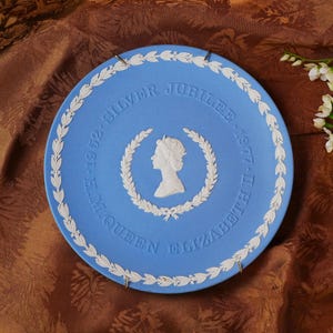 May include: A blue commemorative plate with white lettering and a white silhouette of a person inside a wreath. The plate reads "SILVER JUBILEE 1952-1977 II QUEEN ELIZABETH". The plate has a white floral border and gold hanging hardware.
